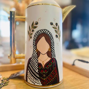 May include: A white ceramic coffee carafe with a gold handle and trim, featuring a hand-painted illustration of a faceless figure wearing a black and white keffiyeh headscarf, a dark patterned garment, a gold key necklace, and gold earrings.