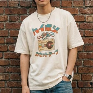 May include: An off-white short-sleeve t-shirt featuring a graphic print of a cassette tape with tangled tape and Japanese text above and below the illustration. The shirt is paired with blue denim jeans and a silver chain necklace.