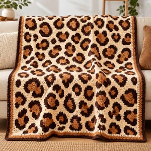 Puede incluir: Una manta de ganchillo rectangular con un patrón repetitivo de leopardo en tonos crema, bronceado y marrón oscuro. La manta tiene un borde sólido de color marrón oscuro que enmarca el diseño animal, con una textura de punto visible.