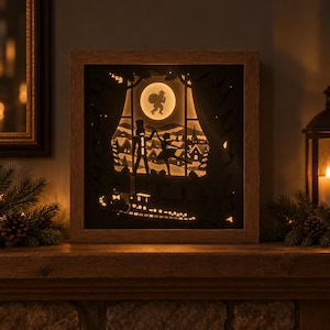 Puede incluir: Una caja de sombras cuadrada de madera con una escena de papel recortado retroiluminada. La silueta muestra a un cascanueces y una bailarina bailando ante una ventana con luna llena, un tren de juguete y un paisaje de pueblo nevado.