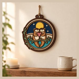 May include: A circular layered wooden wall hanging featuring two brown otters holding a pink heart. The design includes a yellow moon, white stars, and a teal water element, all framed in dark wood with a jute rope hanger at the top for display.