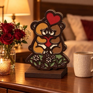 May include: A multi-layered wooden cutout ornament featuring two brown bears embracing and holding a red heart, topped with a red heart shape, standing on a rectangular base, with a green floral design at the bottom of the bear figure.