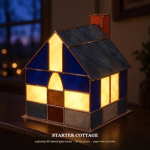 Op de afbeelding: Een oplichtend 3D-glas-in-lood huisje met de tekst STARTER COTTAGE, bestaande uit blauwe, witte en bruine glaspanelen verbonden met koperfolie. De structuur bevat een schoorsteen, een puntdak en verlichte ramen, opgebouwd uit 40 onderdelen.