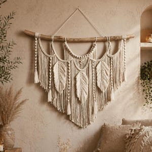 Può includere: Un grande arazzo da parete in macramè color bianco sporco appeso a un ramo di legno naturale. Il design presenta intricati motivi a nodi, tre elementi a frangia a forma di foglia e molteplici cordini pendenti con piccole nappe alle estremità.