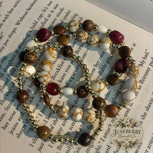 May include: A stack of three beaded bracelets featuring a mix of white and brown marbled beads, dark wood-grain beads, deep red polished stones, and small metallic gold spacer beads. A circular logo with the text JUNEBERRY Bracelet Co. is visible.