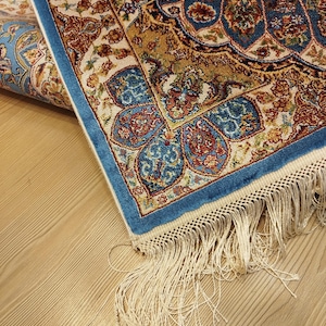 May include: A rectangular area rug featuring a complex floral and geometric pattern in shades of blue, gold, cream, and red. The rug has a distinct blue border and is finished with long, cream-colored fringe tassels along the short edge.