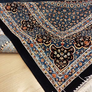 May include: A rectangular prayer rug featuring a deep blue central field and a wide black border. The rug is decorated with intricate floral and vine patterns in shades of light blue, white, orange, and black, finished with white fringe at the edge.