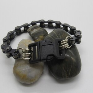 Puede incluir: Un brazalete de cadena negra con un cierre de pl&aacute;stico negro y anillos plateados. El brazalete se muestra sobre dos rocas grises.