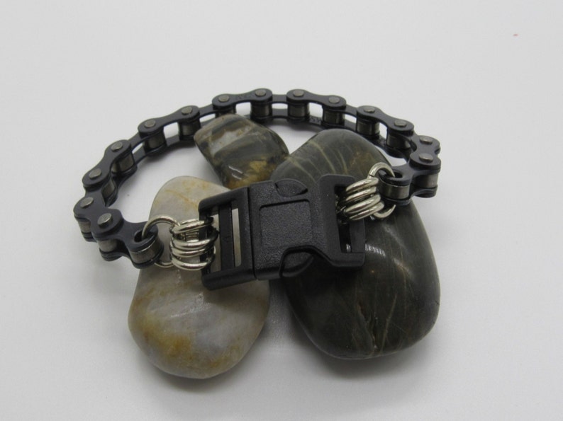 Puede incluir: Un brazalete de cadena negra con un cierre de pl&aacute;stico negro y anillos plateados. El brazalete se muestra sobre dos rocas grises.