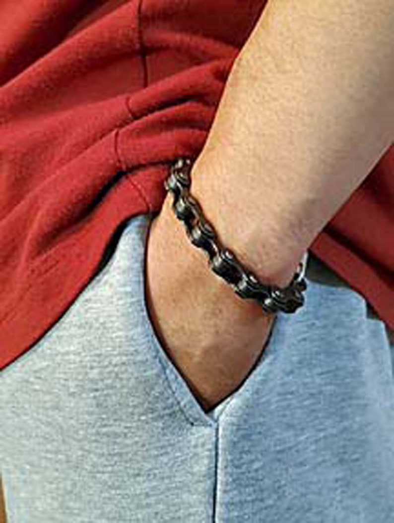 Puede incluir: Un brazalete de cadena de metal negro. El brazalete est&aacute; hecho de piezas de metal enlazadas que se asemejan a una cadena de bicicleta.