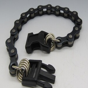 Puede incluir: Un brazalete de cadena negra con dos cierres de pl&aacute;stico negro. La cadena est&aacute; hecha de eslabones met&aacute;licos y los cierres tienen anillos plateados.