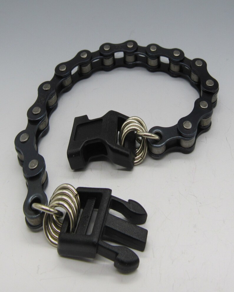 Puede incluir: Un brazalete de cadena negra con dos cierres de pl&aacute;stico negro. La cadena est&aacute; hecha de eslabones met&aacute;licos y los cierres tienen anillos plateados.
