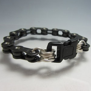 Puede incluir: Un brazalete de cadena de bicicleta negro con un cierre de pl&aacute;stico negro y anillos de metal plateado.