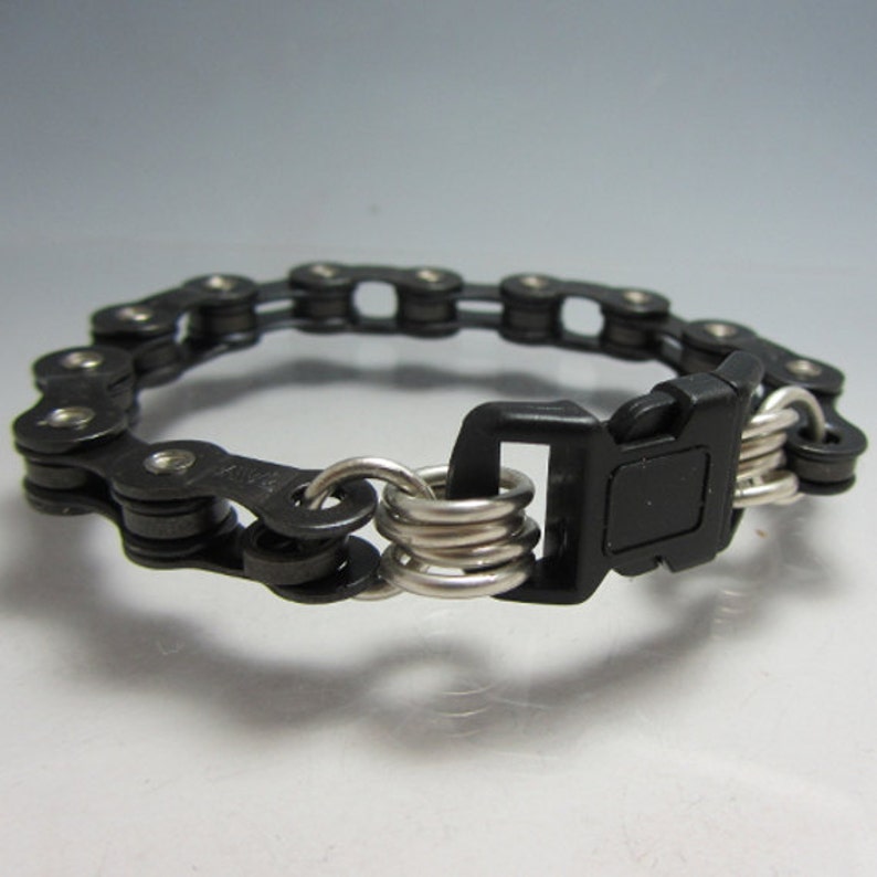 Puede incluir: Un brazalete de cadena de bicicleta negro con un cierre de pl&aacute;stico negro y anillos de metal plateado.