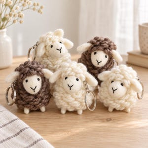 May include: A collection of five small sheep keychains made from textured yarn. Three sheep are cream-colored and two are brown. Each sheep features a smooth, light-colored felt face with embroidered black eyes and a Y-shaped nose and mouth.