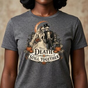 May include: A charcoal gray t-shirt featuring a graphic of two skeletons dressed as a bride and groom sitting on a bench. The text reads Till Death... Still Together, surrounded by pumpkins, a crescent moon, and decorative flourishes.