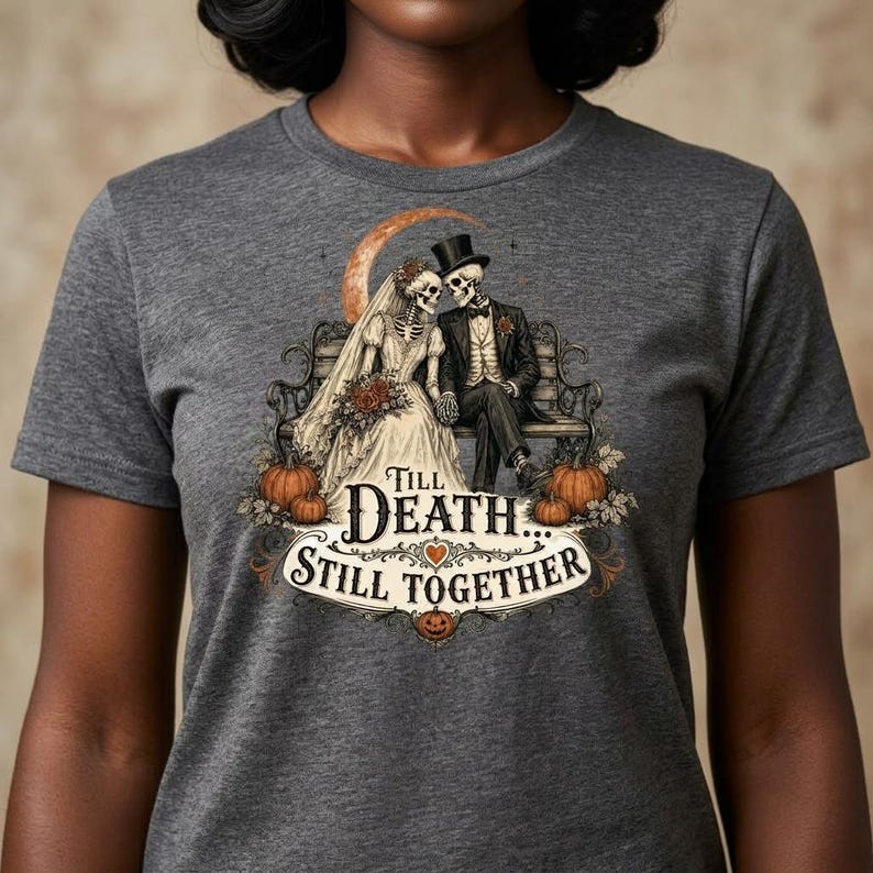 May include: A charcoal gray t-shirt featuring a graphic of two skeletons dressed as a bride and groom sitting on a bench. The text reads Till Death... Still Together, surrounded by pumpkins, a crescent moon, and decorative flourishes.
