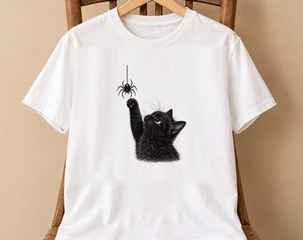 May include: A white short-sleeved t-shirt featuring a graphic illustration of a fluffy black kitten reaching up with one paw to bat at a small black spider hanging from a single thread. The design is centered on the front of the garment.