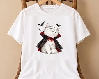May include: A white short-sleeved t-shirt featuring a graphic print of a fluffy white kitten wearing a black cape with a red lining and a red tie. The kitten has its eyes closed and is surrounded by three small black flying bat silhouettes.