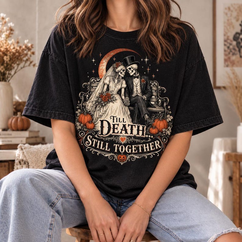 May include: A black t-shirt featuring a graphic of two skeletons in wedding attire sitting on a bench under a crescent moon. The text reads Till Death... Still Together, surrounded by orange pumpkins, red roses, and decorative scrollwork.