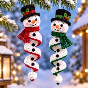 Könnte beinhalten: Zwei gehäkelte Schneemann-Ornamente mit spiralförmigen Körpern. Ein Schneemann trägt einen roten Schal, der andere einen grünen. Beide haben schwarze Zylinder mit Stechpalmen-Akzenten, orangefarbene Karottennasen und schwarze Knöpfe.