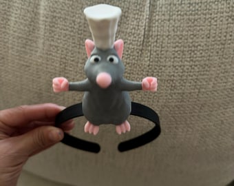 Op de afbeelding: Een zwarte haarband met een grijze ratfiguur die een witte koksmuts draagt. De rat heeft roze oren, een roze neus, roze handen en roze voeten, met de armen wijd uitgestrekt. Het figuurtje is gecentreerd op de bovenkant van de dunne haarband.