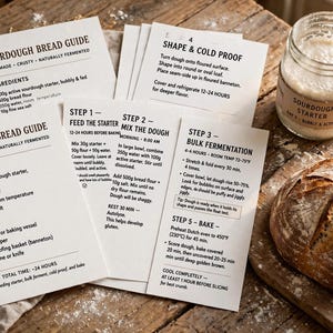 May include: A set of white instructional cards titled Sourdough Bread Guide with black text detailing ingredients, equipment, and steps. Beside the cards sits a glass jar of sourdough starter and a round, crusty loaf of baked bread.