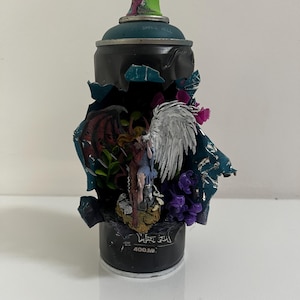 Könnte beinhalten: Eine schwarze Sprühdose mit einem großen Ausschnitt, der ein Miniatur-Diorama im Inneren enthüllt. Das Diorama zeigt eine Figur mit einem dunkelroten und einem weißen Flügel, umgeben von kleinen violetten Blumen und grünem Blattwerk. Die Dose trägt den Text 400.M.
