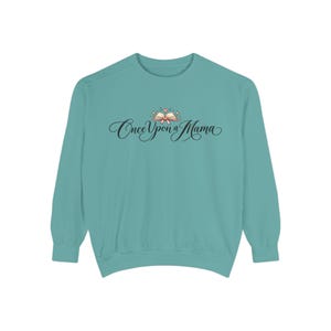 May include: A teal long-sleeve crewneck sweatshirt featuring the black cursive text Once Upon a Mama centred on the chest. Above the text is a small illustration of an open book with a pink ribbon and tiny sparkling star accents.