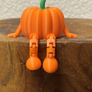 May include: A 3D printed orange pumpkin figurine with a green stem, featuring articulated arms and dangling legs that allow it to sit on an edge. The pumpkin has a ribbed texture and rounded feet, showcasing a smooth, matte plastic finish.