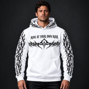 May include: A white hooded sweatshirt featuring black tribal-style graphic patterns on the sleeves and hood. The chest displays a black graphic design with the text "RIDE AT YOUR OWN RISK" printed in a stylised, sharp-edged black font.