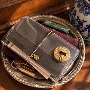 May include: A dark-colored notebook encased in a clear plastic cover, secured with an elastic band and a gold-toned circular keyhole charm. A small gold-toned metal charm and a brass-colored cylindrical object are attached to the elastic band.