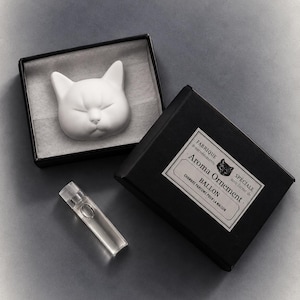 Puede incluir: Un adorno aromático de cerámica blanca con forma de gato en una caja negra con un pequeño frasco de vidrio transparente. La etiqueta de la caja dice: FABRIQUE de eter méditerranis, Aroma Ornement, BALLON, SPÉCIALE sous forme de, CHAMBRE PARFUMS POUR LA MAISON.