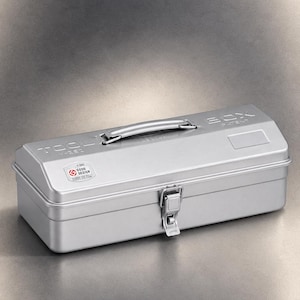 May include: A silver-colored metal tool box with a hinged lid, a central handle, and a front latch. The lid features embossed text reading TOOL BOX and Y-350, along with a Good Design sticker. The word JAPAN is printed on the front latch.