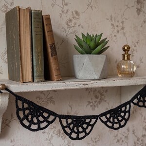May include: A black crocheted garland featuring a repeating web-like pattern suspended from two brass-colored knobs on a white wooden shelf. The shelf holds three vintage hardcover books, a small green succulent in a geometric pot, and a glass bottle.