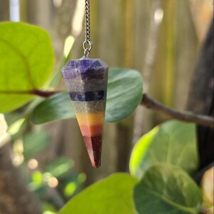 May include: A faceted, cone-shaped pendulum made of stacked, multi-colored stone segments including purple, gray, blue, green, yellow, orange, and red, suspended from a thin silver-toned metal chain attached to a small metal loop at the top.