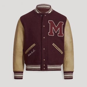 Könnte beinhalten: Eine Collegejacke mit einem dunkelburgunderroten Wollkörper und beigefarbenen Lederärmeln. Die Vorderseite zeigt ein großes burgunderrotes M mit weißem Rand, Metalldruckknöpfe und den gestickten Schriftzug Polo R.L. im Bereich der rechten Tasche.