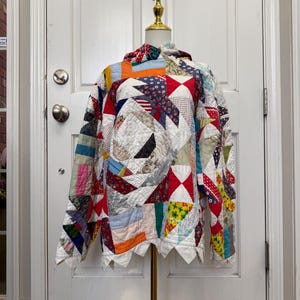 May include: A long-sleeve pullover top constructed from a patchwork quilt featuring a variety of colorful fabric scraps in red, blue, orange, yellow, and white. The garment has a hood and a jagged, pointed hemline along the bottom edge.