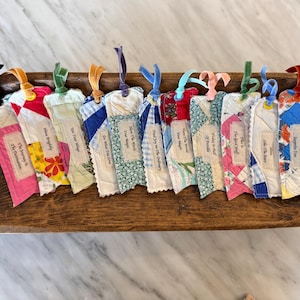 Può includere: Una collezione di undici segnalibri rettangolari in tessuto realizzati con ritagli di patchwork trapuntato in vari colori e fantasie. Ogni segnalibro presenta una piccola etichetta bianca con testo e un nastro di velluto colorato in cima.
