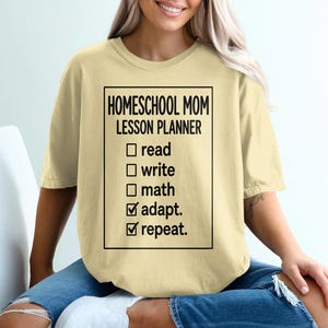 May include: A light yellow short-sleeve t-shirt featuring a black rectangular graphic with the text HOMESCHOOL MOM LESSON PLANNER followed by a checklist with the items read, write, math, adapt, and repeat, with the last two items checked.