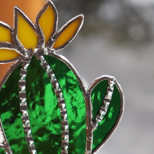 May include: A stained glass cactus sculpture with a yellow flower and green leaves, set in a white ceramic pot filled with small pebbles. The cactus is on a wooden coaster.