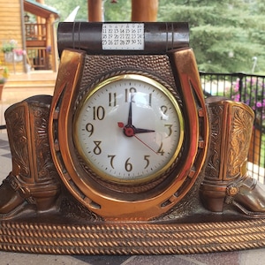 May include: A bronze-colored clock with a horseshoe design, featuring a clock face with black numerals and red hands. The clock has a calendar and is flanked by two cowboy boot sculptures. The base has a rope-like texture.