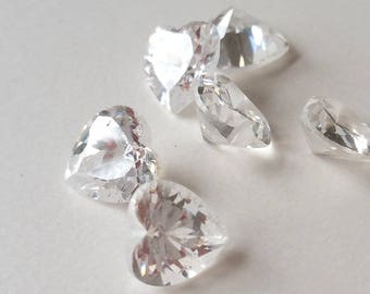 May include: A group of six clear heart-shaped crystal rhinestones. The rhinestones are scattered on a white surface.