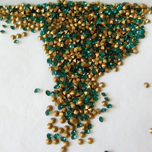 May include: A large quantity of small, green and gold rhinestones. The rhinestones are scattered on a white surface.