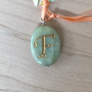 May include: A green stone pendant with a gold letter F engraved on it. The pendant is attached to a peach-colored ribbon.