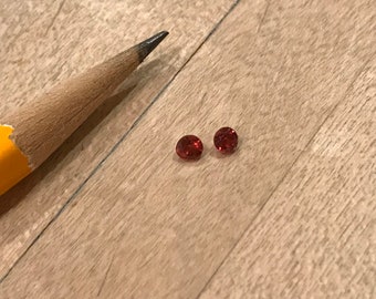 May include: Two small, round, red gemstones on a wooden surface. A yellow pencil is in the background.