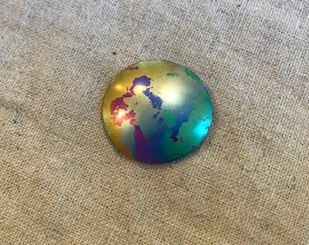 May include: A round, metallic button with a gold base and a colorful, abstract design. The design features splashes of blue, green, purple, and red.