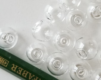 May include: A collection of clear glass beads, some with a slight iridescent sheen, scattered on a white surface. A green pencil with gold lettering is partially visible in the bottom right corner.