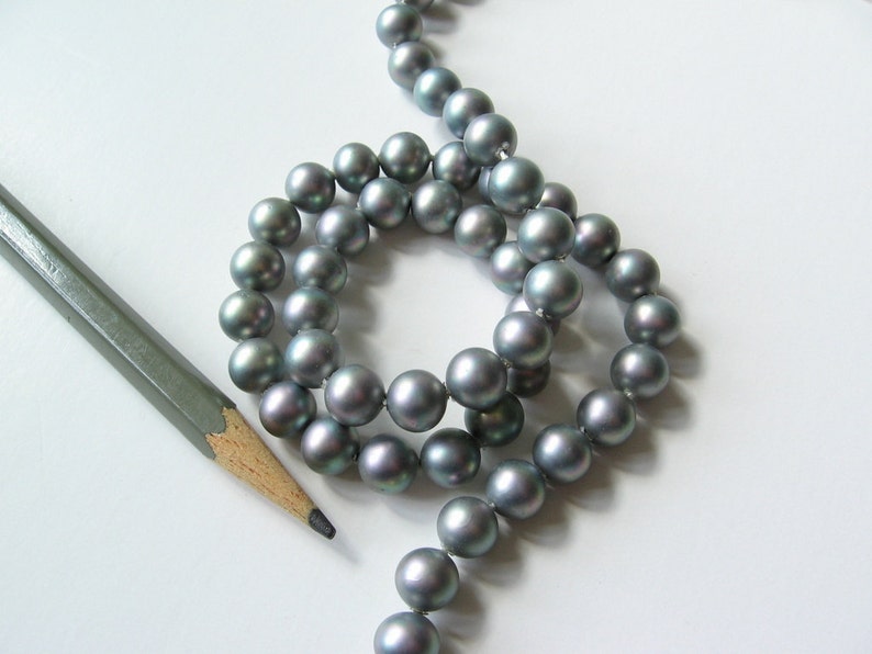 May include: A strand of gray pearls, arranged in a circle, with a pencil lying on a white surface.
