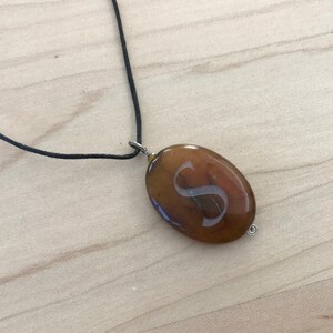 May include: A brown oval pendant necklace with a silver letter 'S' engraved on it. The necklace is made with a black cord.